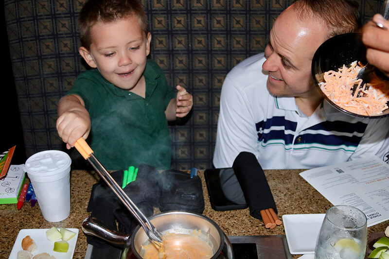 A young boy stirring cheese fondu as his dad watches
