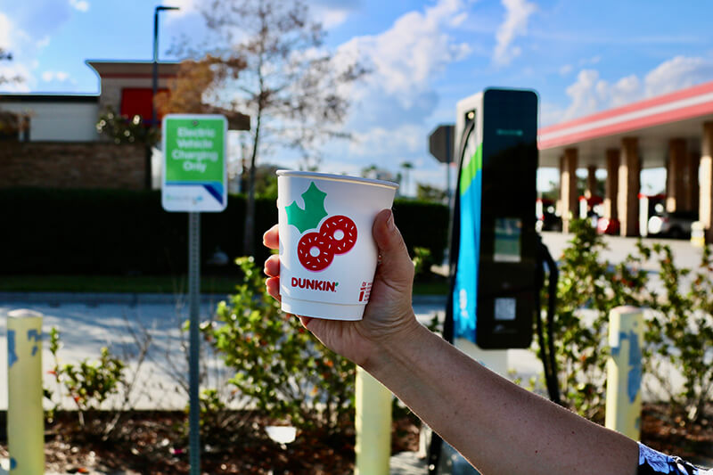 Hand holding up a cup of Dunkin' beverage in front of an EV charging station
